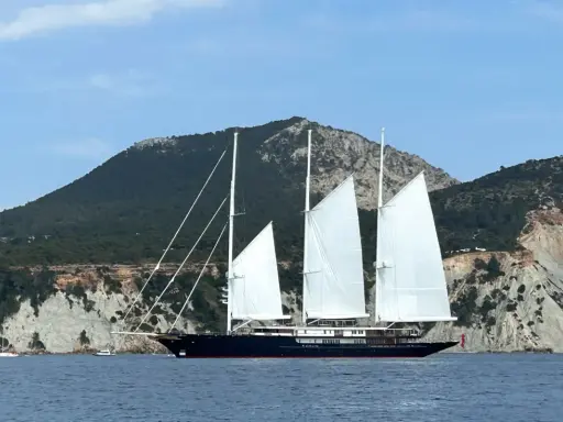 Le yacht de Bezos, un voilier majestueux, navigue en Méditerranée, ses voiles blanches déployées devant des falaises escarpées.