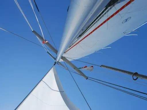 Vue plongeante sur le mât et les voiles d'un voilier, le ciel bleu intense en arrière-plan. L'image capture l'essence de la navigation, le vent gonflant les voiles.