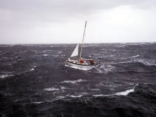 Un voilier blanc lutte contre les vagues déchaînées dans une mer agitée. Le ciel est gris et menaçant, annonçant une tempête.