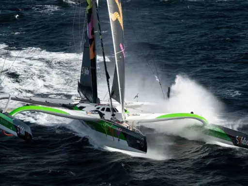 Le trimaran Sodebo, un bateau ultime, fend les vagues sombres, projetant des gerbes d'eau dans une course effrénée.