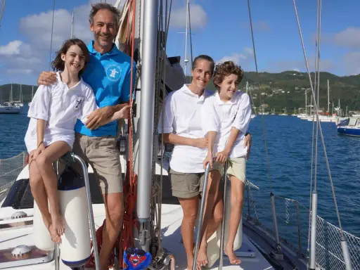 Famille souriante sur un voilier, un moment clé de leur récit de voyage en voilier. La mer bleue et les voiliers en arrière-plan complètent ce tableau idyllique.