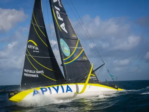 Le voilier Apivia, skippé par Alex Thomson, fend les vagues sous un ciel bleu et nuageux.