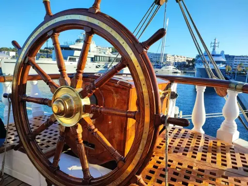 Roue de gouvernail en bois et laiton sur le pont d'un bateau. En arrière-plan, des yachts modernes et des bâtiments dans un port ensoleillé.