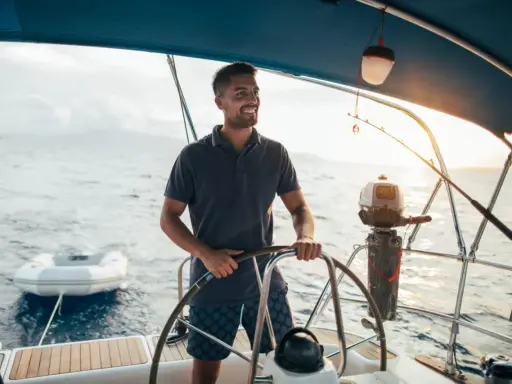 Un homme souriant barre un bateau, profitant du soleil couchant. Un petit bateau gonflable est attaché à l'arrière.