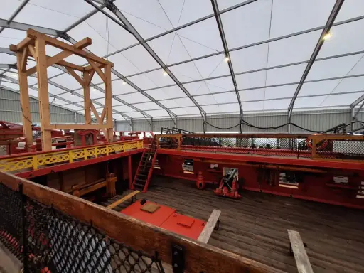 L'intérieur du **bateau Hermione** est en cours de restauration. On voit la structure en bois, les ponts rouges et jaunes, et un canon.