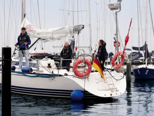 Trois personnes sur un bateau, prêtes à appareiller. Le drapeau allemand flotte fièrement. L'erre bateau est calme.
