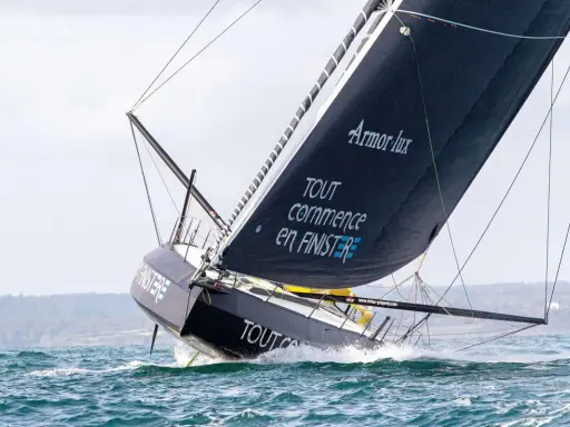 Le voilier IMOCA de Jean Le Cam fend les vagues, sa grand-voile noire ornée du slogan "Tout commence en Finistère" gonflée par le vent.