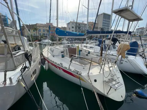 Un voilier First 32, amarré dans un port, avec d'autres bateaux et des bâtiments en arrière-plan.