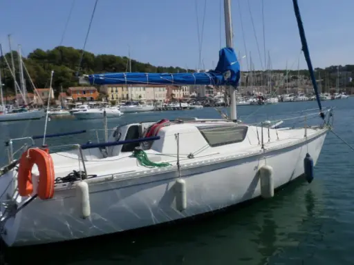 Bateau blanc à quai, voiles bleues enroulées. Au loin, un port animé avec des maisons colorées et d'autres voiliers. Un endroit idéal pour un séjour, comme le suggèrent les gib sea 76 avis.