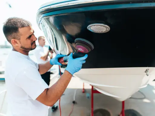 Un homme polit la coque d'un bateau. Il utilise une polisseuse pour un travail de précision, peut-être en prévision d'une révision moteur inboard.