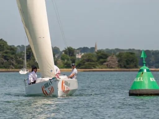 Voile blanche sur l'eau, le voilier "Bihan 6.50" navigue avec deux personnes à bord. Une bouée verte marque le passage.