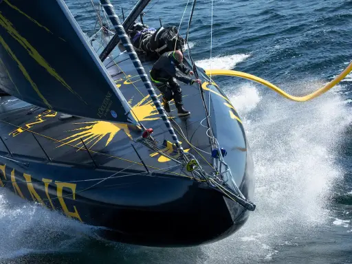 Un skipper manœuvre sur l'IMOCA Bureau Vallée, le bateau noir et jaune fend les vagues dans un embrun spectaculaire.