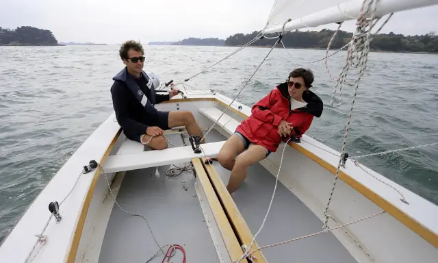 Deux personnes profitent d'une journée ensoleillée sur un voilier dayboat, naviguant sur une mer calme.