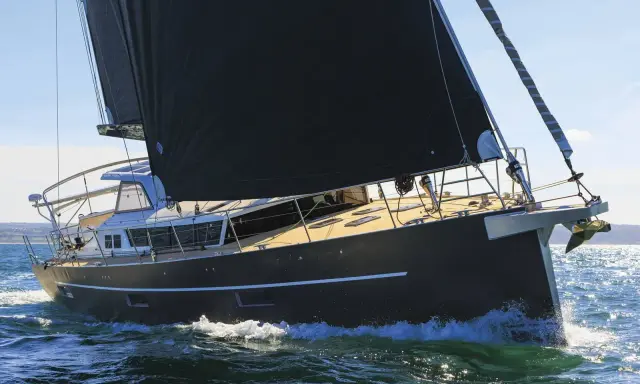 Un voilier moderne, avec une grande voile noire, navigue sur une mer agitée. Xavier Desmarest admire la vue depuis le pont.