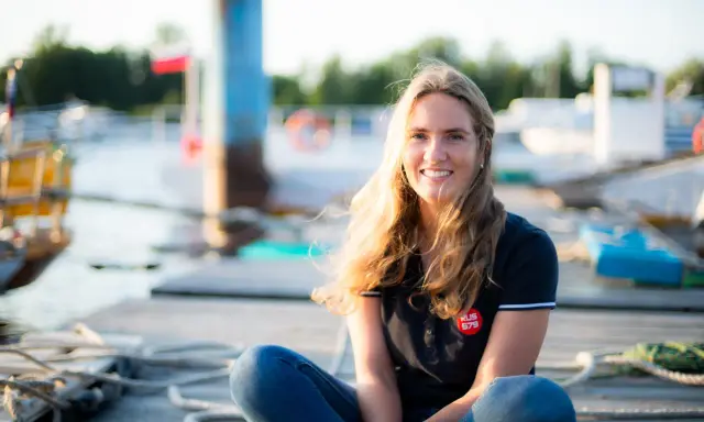 Irina Gracheva, souriante, assise sur un ponton en bois, avec des bateaux et des bouées en arrière-plan.