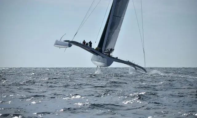 Un maxi trimaran fend les vagues, ses coques latérales hors de l'eau, propulsé par une voile immense. L'équipage est visible sur le pont.
