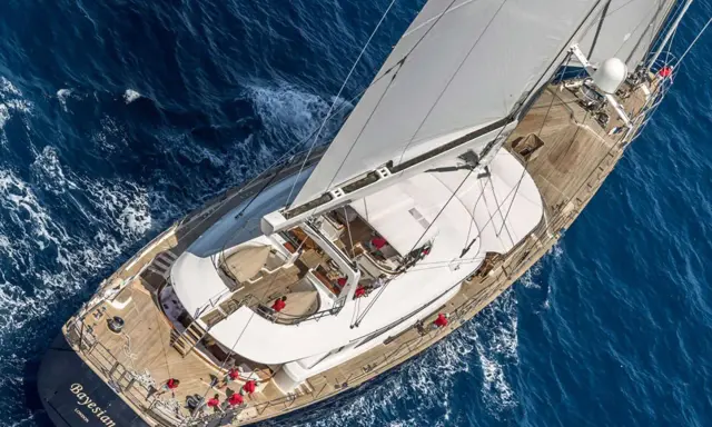 Le yacht "Bayesian" navigue sur des eaux bleues, ses voiles blanches gonflées par le vent. L'équipage en rouge s'affaire sur le pont en bois.