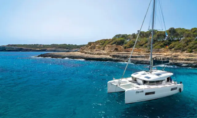 Un catamaran blanc, le "StarElli", navigue dans des eaux turquoise près d'une côte rocheuse et verdoyante. Ce safran catamaran offre une vue idyllique.