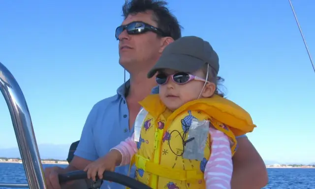Un homme, le chef de bord bateau, tient son enfant sur ses genoux, naviguant sous un ciel bleu.