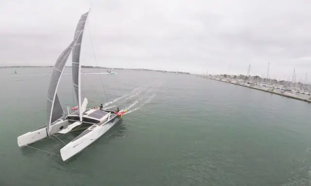 Un catamaran TS3 navigue sur une mer calme, avec un port rempli de voiliers à droite.
