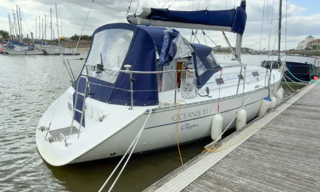 Un voilier blanc, un Océanis 311 Clipper, est amarré à un quai en bois. Des pare-battages blancs protègent sa coque.