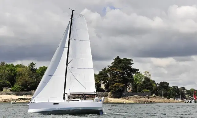 Voilier blanc moderne naviguant sur l'eau, avec des avis sur le Pogo 30. Paysage côtier verdoyant en arrière-plan.