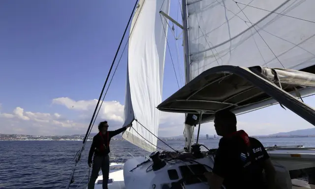 Deux hommes sur un catamaran, l'un ajuste la voile pour se mettre à la cape, tandis que l'autre tient la barre. Le soleil brille sur la mer bleue.