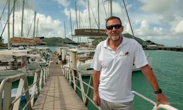 Loïc Bonnet, un homme souriant, pose sur un ponton bordé de voiliers. Il porte un polo Dream Yacht Charter, prêt pour une aventure en mer.