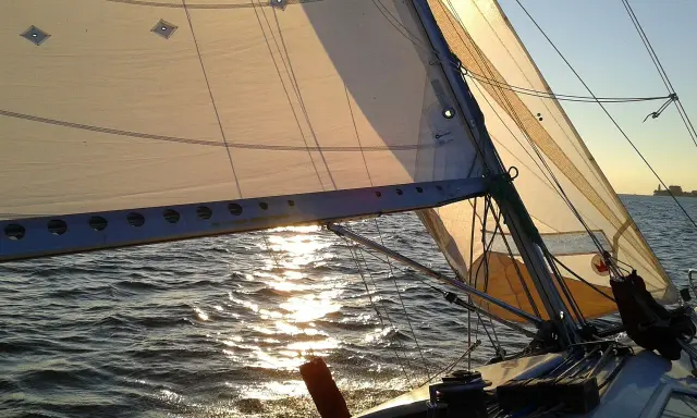 Voile d'un bateau au coucher du soleil, reflets dorés sur l'eau. La balancine du bateau est bien tendue.