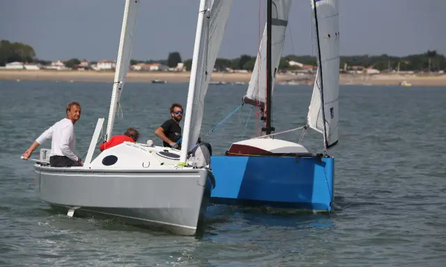 Deux voiliers naviguent côte à côte. Sur le premier, un homme en blanc manœuvre. Le second, d'un bleu vif, est un aloes 18.