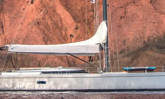 Un voilier Swan 76, avec une voile blanche déployée, navigue près d'une falaise rouge. Un kayak est rangé sur le pont.