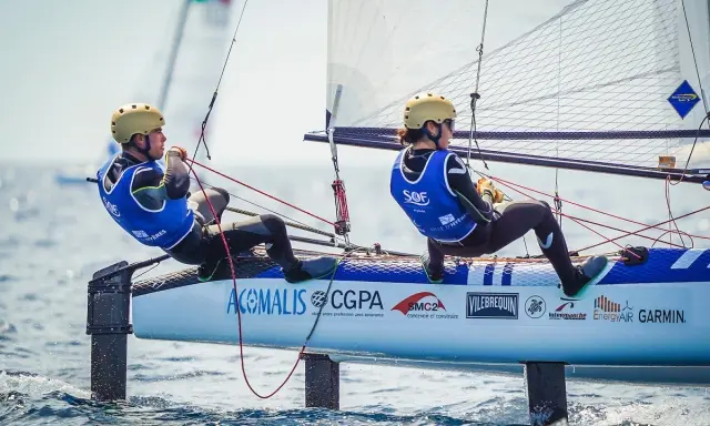 Deux jeunes navigateurs, dont Lou Berthomieu, en action sur un catamaran de sport, le vent gonflant les voiles.