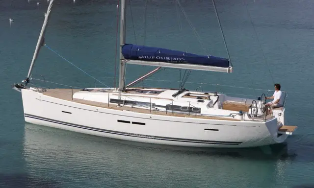 Un voilier blanc Dufour 405 Grand Large navigue sur une eau turquoise. Un homme est au poste de barre.