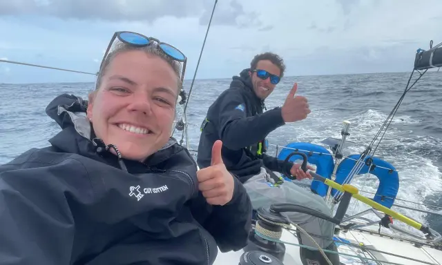 Hugo Dhallenne et sa coéquipière souriants sur un voilier, pouces levés, en pleine mer sous un ciel couvert.