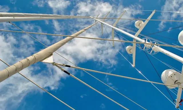 Vue plongeante sur le gréement d'un bateau, mâts blancs et cordages fins se découpant sur un ciel bleu et nuageux.