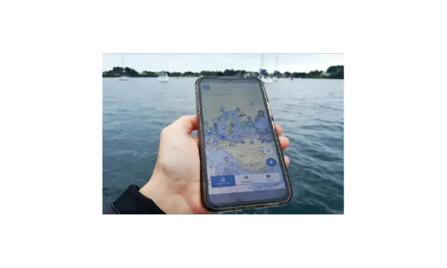 Main d'une personne tenant un smartphone affichant une carte marine avec des points d'intérêt. Le nav et co. sont visibles sur l'eau.