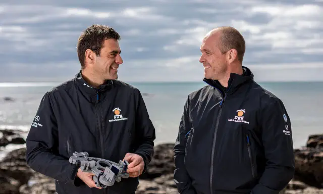Damien Guillou et son collègue souriants sur la plage, l'un tenant un sextant, prêts pour une nouvelle aventure maritime.