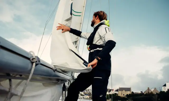 Un homme ajuste un type de voile sur un voilier, avec des maisons en arrière-plan.