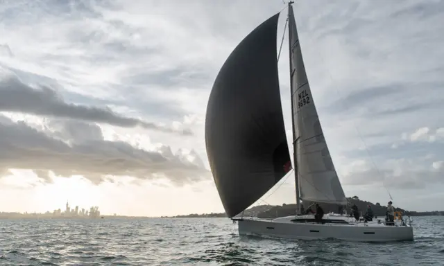 Un voilier navigue avec un grand gennaker noir gonflé par le vent, contrastant avec le spinnaker gris. La ville d'Auckland se dessine à l'horizon.