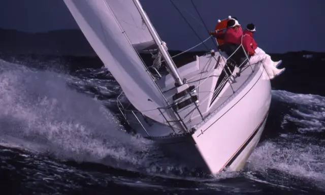 Voilier blanc fendant les vagues, deux personnes en ciré rouge. Une scène de navigation intense, digne des premiers 285 avis.