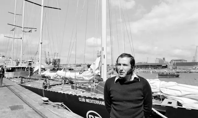 Un homme pose devant le voilier "Club Mediterranee". Ce navigateur célèbre semble prêt pour une nouvelle aventure maritime.