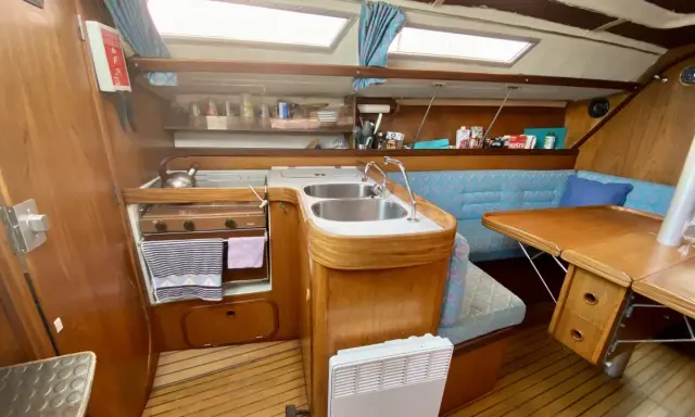 Cuisine de voilier avec cuisinière, évier double, banquette bleue et table en bois. Un radiateur est visible. 312 avis gib sea.