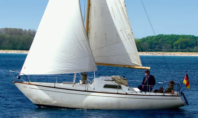 Un voilier blanc navigue sur une mer bleue, ses voiles gonflées par le vent. L'écume de mer danse autour de la coque. Deux personnes et un chien profitent de la balade.