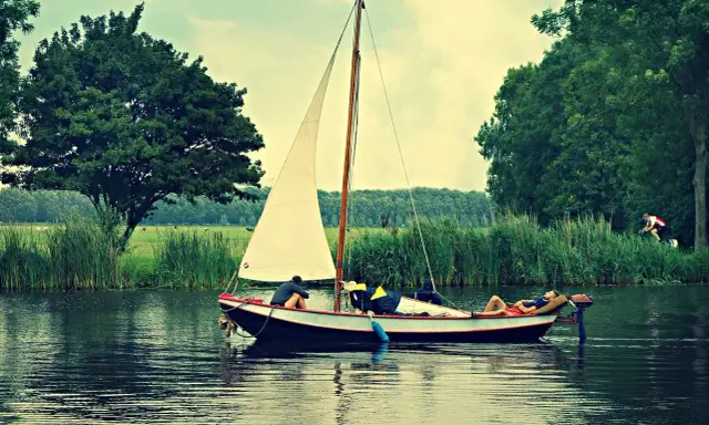 Un petit type de bateau à voile navigue sur un canal bordé d'arbres. Une personne se repose sur le pont, une autre est assise.