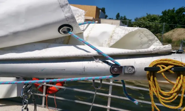 Le cordage bleu passe dans un œillet de la voile, prêt à ajuster la bosse de ris. Une amarre jaune est enroulée sur un taquet.