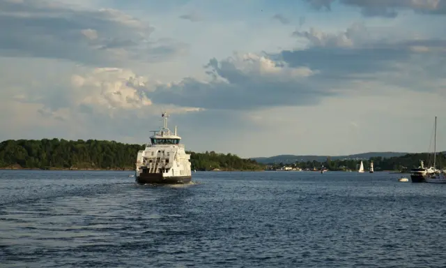 Un ferry blanc navigue sur une eau bleue, laissant une traînée derrière lui. La vitesse du bateau est visible dans le sillage. Au loin, des voiliers et une côte boisée.