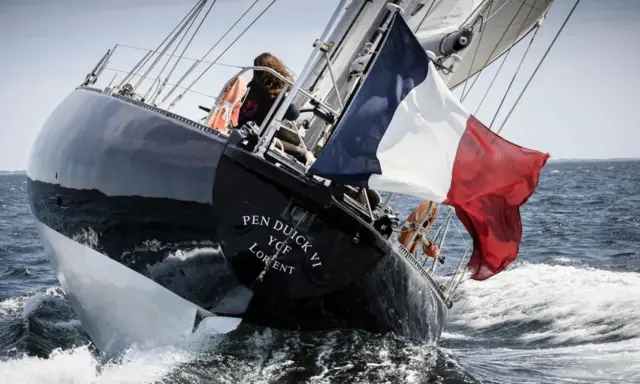 Le voilier Pen Duick VI fend les vagues, pavillon français au vent. Une silhouette se tient à la barre.