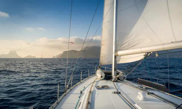 Le voilier navigue sur une mer bleue, le grand voile blanc gonflé par le vent. Au loin, des îles brumeuses se dessinent sous un ciel clair.