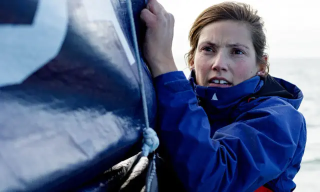 Tiphaine Ragueneau, concentrée, ajuste une voile bleue sur un bateau. La mer est visible en arrière-plan.