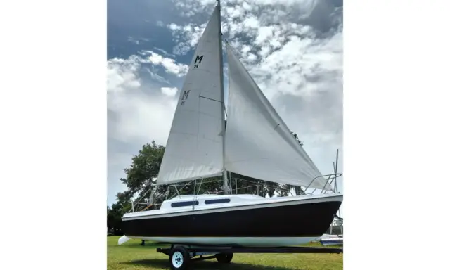 Voilier Mac Gregor 25, prêt à naviguer, avec ses voiles blanches gonflées par le vent sous un ciel nuageux.